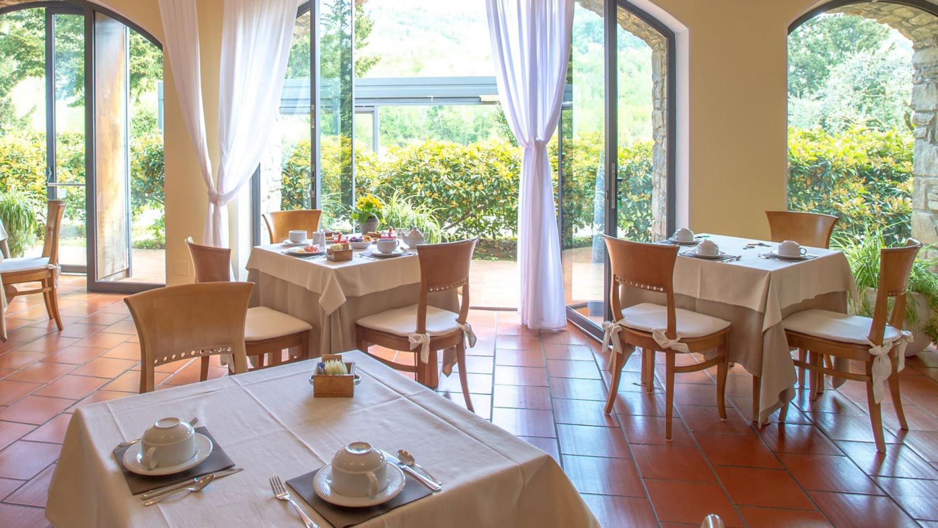 Ristorante luminoso con tavoli apparecchiati, vista giardino e tende bianche eleganti.