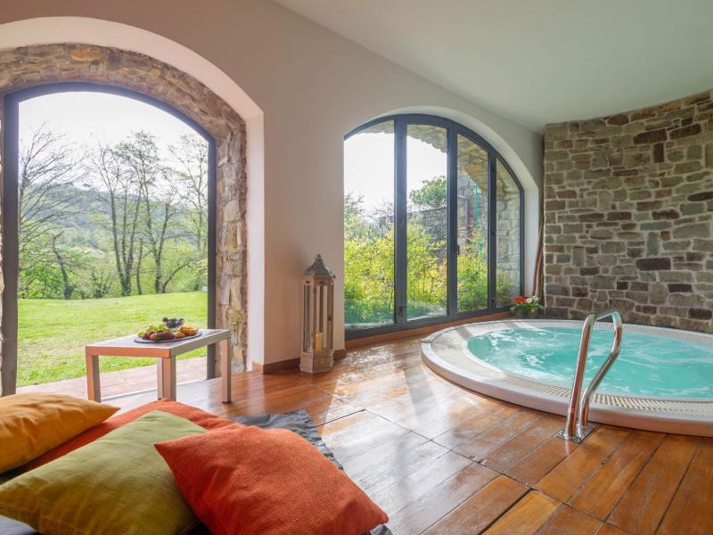 Elegante spa con jacuzzi, vista sul giardino, cuscini colorati e tavolino con frutta.
