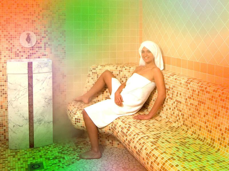 Donna rilassata in sauna, avvolta in asciugamano, seduta su panchina piastrellata.