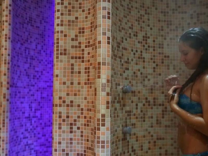 Donna sotto la doccia con piastrelle colorate e luce viola sullo sfondo.